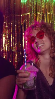 Kıvırcık kızıl saçlı ve kalp şeklinde güneş gözlüklü genç bir kadın altın sarısı bir partide kokteyl içiyor.