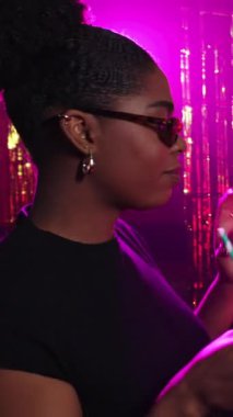 Neşeli bir gece hayatı yaşayan kendine güvenen genç bir kadın, renkli disko ışıkları altında canlı enerjisiyle kokteyl yudumluyor ve dans ediyor.