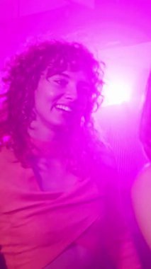 Hareket eden ve müzikle oyalanan kadın arkadaşlar, canlı, yavaş çekim gece kulübü atmosferinde dans ederken neşe saçan renkli ışıklar ve enerjik ritimle