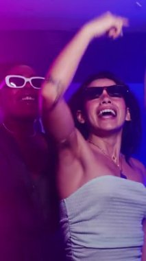 Enerjik genç arkadaşlar, son moda güneş gözlüğü takıp birlikte dans ediyorlar, neon ışıkları ve neşeli atmosferi olan canlı bir gece kulübünde müzik dinliyorlar.