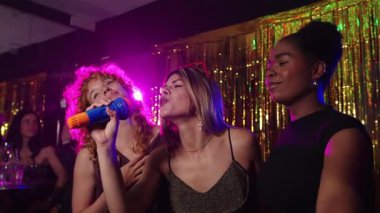 Bir grup farklı bayan arkadaş tutkuyla karaoke söylüyor, neon ışıklarıyla çevrili altın sahnedeki mikrofonlarla neşeli bir müzik anı paylaşıyorlar.