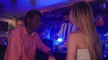 Genç çift barmenle etkileşime giriyor, samimi bir anı paylaşıyor ve loş ışıklı şehir barında mavi ışıklandırma ve şık bir ortam ile renkli kokteyller ısmarlıyor.