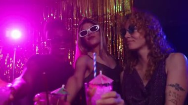 Şık, çok ırklı kadınlar ve erkek, gece kulübünde canlı mor ışık ve altın fon eşliğinde içki içiyorlar.