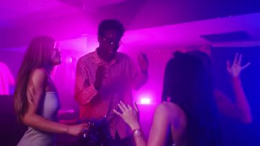 Enerjik hareketlerle dans eden enerjik genç yetişkinler, gece kulübünde titreşimli müziğin ve renkli neon ışıkların tadını çıkarıyorlar.