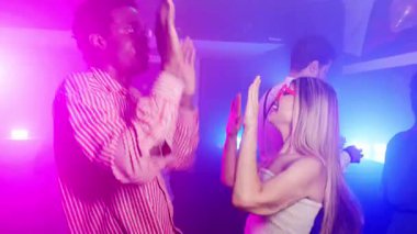 Enerjik çok ırklı çift neşeli dans anlarını paylaşıyor, el çırpıyor ve yavaş çekim kutlamalarında canlı gece kulübü ışıkları altında ritmik bir şekilde hareket ediyorlar.