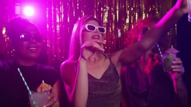 Hayat dolu gece hayatının tadını çıkaran, renkli kokteyller içen ve altın tonlu yavaş çekim arkaplanlı görkemli disko partisinde dans eden kadın arkadaşlar.