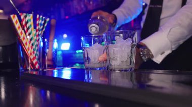 Profesyonel barmen alkollü içeceği gece kulübü ortamında buzla dolu iki bardağa dolduruyor.