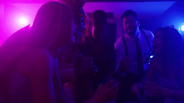 Çok ırklı arkadaşlar gece hayatının tadını çıkarıyorlar, renkli neon ışıklar altında yavaş çekimde içkilere kadeh kaldırıyorlar.
