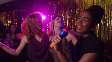 Bir grup bayan arkadaş enerjik bir şekilde birlikte şarkı söylüyor, canlı bir gece kulübünde mikrofonlarla karaoke yapıyor, kızlar gecesi yavaş çekimde müzik dinlemenin keyfini çıkarıyorlar.