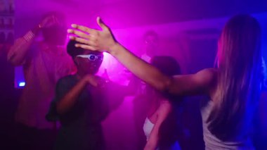 Kalabalık gece kulübü dans pistinde dans eden enerjik, çok ırklı arkadaş grubu, yavaş çekimde renkli neon ışıkların altında heyecanla hareket ediyorlar.