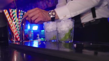Profesyonel barmen mojito karıştırıyor, özenle malzemeler ekliyor ve nane şekerini karıştırıyor, limon ezilmiş buzla kaygan bar tezgahında canlı ve yavaş çekimde ışıklandırılıyor.