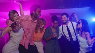 Çok kültürlü genç arkadaşlar enerjik bir şekilde dans ediyor, neon renkli ışıklandırmayla gece hayatının tadını çıkarırken içkileri paylaşıyor ve hareketli sosyal anlar yaratıyorlar.