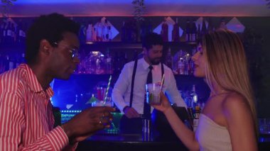 Genç çift samimi bir anı paylaşıyor şık şehir barında renkli kokteyller yudumluyor canlı bir akşamda, dinamik yavaş çekim sekansıyla romantik bağları yakalıyor.