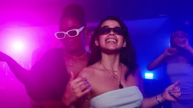 Modayı takip eden güneş gözlüğü takan kadınlar kalabalık gece kulübü dans pistinde arkadaşlarıyla dans ediyor, gece hayatı kutlamaları sırasında canlı bir sosyal enerji yaratıyorlar.