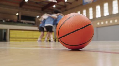 Turuncu basketbol topu, arka planda birbirini tebrik eden bayan oyuncuların olduğu ahşap bir sahada yatıyor.