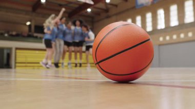 Turuncu basketbol, arka plandaki zaferi kutlayan bayan lise ya da üniversite basketbol takımı gibi ahşap sahada yatar.