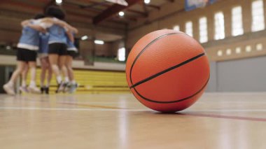 Turuncu basketbol sahada yatarken, bayan basketbol takımı arka planda zaferi kutluyor.