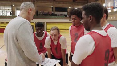 Basketbol koçu beyaz tahtaya stratejik oyun çiziyor, kapalı sahadaki yoğun takım toplantısında motive olmuş atletlere rehberlik ediyor.