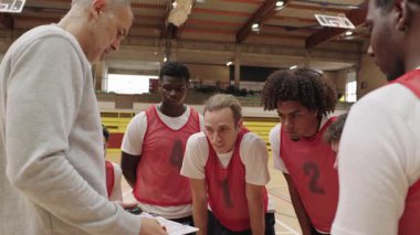 Basketbol koçu stratejik toplanma sırasında takımın yanında duruyor, oyun planını analiz ediyor ve sporculara odaklanmış antrenman boyunca rehberlik ediyor.