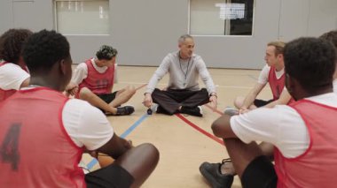Basketbol takımı koçla toplanıyor, spor salonundaki yoğun antrenman sırasında dikkatle dinliyor ve stratejilerini tartışıyor.