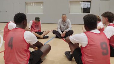 Genç basketbol oyuncularından oluşan bir takım toplanıyor, antrenmandan önce koçların stratejik rehberliklerini dinliyorlar, takım çalışması ve motivasyonlarını gösteriyorlar.