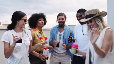 Çok ırklı genç arkadaşlar neşe dolu anları paylaşıyor, içki içerken gülüyor ve kaynaşıyor çatıdaki sosyal toplanma sırasında panoramik şehir manzaralarının keyfini çıkarıyorlar.