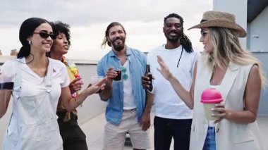 Çeşitli arkadaş grupları şehir terasında dinleniyor, sıcak yaz akşamı günbatımında içkileri ve kahkahaları paylaşıyorlar.