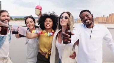 Çeşitli genç arkadaşlar içki paylaşıyor, kadeh tokuşturuyor ve güneşli terasta panoramik şehir manzarasının keyfini çıkarırken neşeyle kutluyorlar.