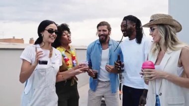 Çeşitli genç arkadaşlar günbatımında kokteyllerini yudumlayarak gülüyor, canlı şehir manzarası ile çatıda buluşuyorlar.