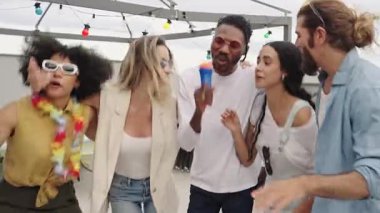 Çok ırklı arkadaşlar çatıdaki karaoke sırasında neşeli anları paylaşıyorlar, güneşli gökyüzünün altında bulaşıcı enerjiyle şarkı söyleyip dans ediyorlar.