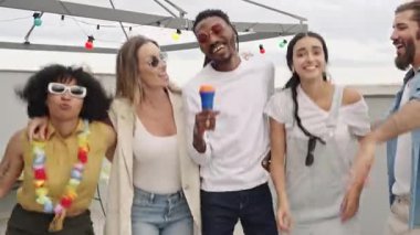 Neşeli çoklu etnik arkadaşlar karaoke yapıyor, dans ederken canlı müzik eşliğinde kaynaşıyorlar ve yaz partisinde panoramik şehir ışıklarının tadını çıkarıyorlar.