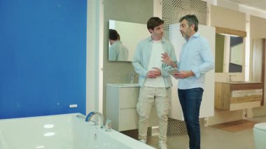 Profesyonel pazarlamacı müşteriye banyo malzemeleri seçimi konusunda rehberlik ediyor, modern sergi ortamında detaylı ürün sunumu için tablet kullanılıyor