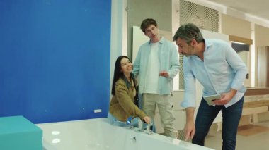 Modern banyo armatürünü seçerken, genç çift beyaz küvete göz gezdirirken satıcı mavi duvarın yanında dijital tablet kullanarak ayrıntıları açıklıyor.
