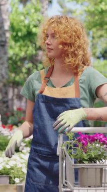Bahçıvan olarak çalışan genç bir kadın çiçek topluyor ve onları yavaş çekimde bir alışveriş arabasına koyuyor.