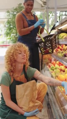 Market çalışanı, temiz ve iyi stoklanmış pazarda müşteriye hizmet ederken renkli ürünlerden olgun, organik hindistan cevizi seçerken dikkatle eldiven giyiyor.