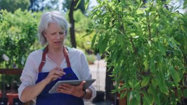 Profesyonel kadın bahçıvan dijital tablet üzerinde bitki verilerini analiz ederken çocuk odasında durup güneşli çalışma gününde yenilikçi teknolojiyle detaylı botanik araştırmaları yakalıyor.