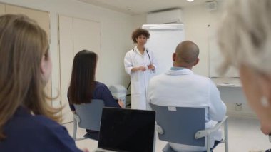 Kadın doktor, meslektaşlarına yavaş çekimde konseptleri anlatırken el kol hareketi yaparak tıbbi eğitim oturumunu yönetiyor.