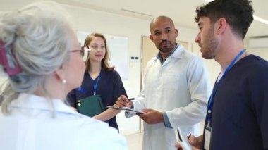 Tıbbi uzmanlar iş birliği yapıyor, hasta verilerini gözden geçiriyorlar, aynı zamanda parlak hastane koridorunda duruyorlar, profesyonel ekip sağlık bilgilerini yavaş çekimde analiz ediyor.