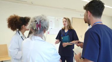 Tıbbi uzmanlar hasta detaylarını tartışıyor, ağır çekimde hastane koridorlarında görüşlerini paylaşıyor ve odaklanmış yoğunlukla işbirliği yapıyorlar.