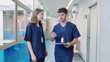 İki genç doktor hastane ortamında hasta bakımıyla ilgili bir tartışmada tıbbi kayıtları inceliyor.
