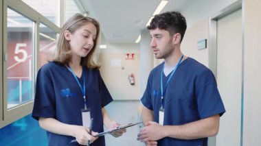 Önlüklü iki sağlık uzmanı ağır çekimde hastane koridorunda sağlık raporlarını inceledi ve tartıştı.