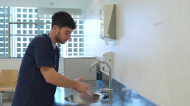 Genç erkek hemşire ellerini titizlikle bir hastane lavabosunda yıkıyor, yavaş çekimde hijyen ve güvenliği vurguluyor.