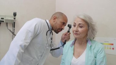 Experienced physician performing detailed ear examination on elderly patient using otoscope during comprehensive health checkup in modern medical clinic setting