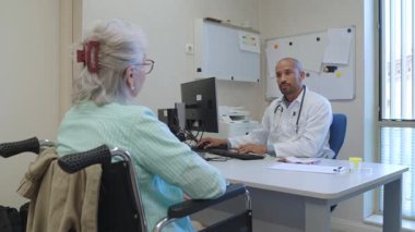 Klinikteki detaylı sağlık muayenesi sırasında tekerlekli sandalyedeki yaşlı kadın hasta tıbbi uzmanlara danışarak, özenli iletişim yoluyla tıbbi görüşleri paylaşıyor.