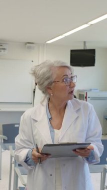 Beyaz önlüklü kıdemli kadın doktor elinde dosya tutarken tıbbi uzmanlığını paylaşıyor, yavaş çekimde klinik danışmanlık sırasında profesyonel sağlık bilgilerini iletiyor.