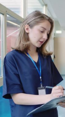 Hasta bilgilerini panoya kaydeden profesyonel hemşire, hastane koridorlarında ağır çekimde yürüyen, beyaz üniformalı.