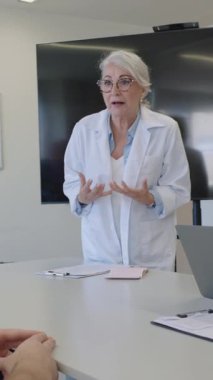 Bir tıp konferansında konuşan kıdemli kadın doktor, araştırma bulgularını sunuyor ve modern bir hastane ortamında iş arkadaşlarıyla görüşüyor.