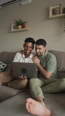 Eşcinsel çift birlikte rahatlıyor, oturma odasındaki kanepede dizüstü bilgisayar kullanıyor, özel bir bağ ve rahatlık anı paylaşıyorlar.