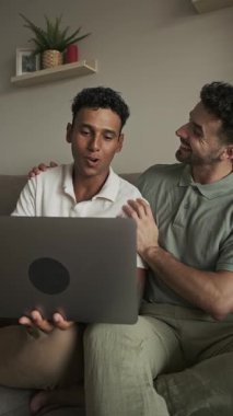 Neşeli eşcinsel partnerler eğlenceli anları paylaşıyorlar, birlikte gülüyorlar, dizüstü bilgisayarın içeriğini izlerken rahat oturma odası ortamında, samimi bir bağ kuruyorlar.