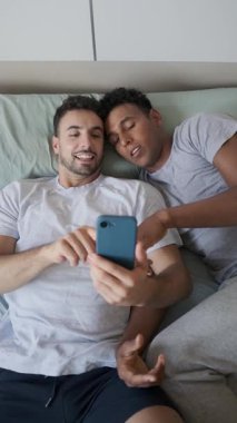 Mutlu lgbti çifti yatakta uzanıyor, akıllı telefon içeriğine bakarken bir anlık neşe paylaşıyorlar, yavaş çekimde çekiyorlar.